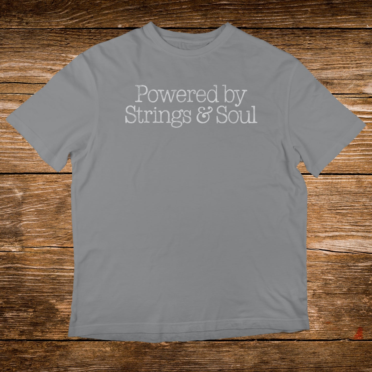 Grey t-shirt with "Powered by Strings & Soul" text on a wooden background.
