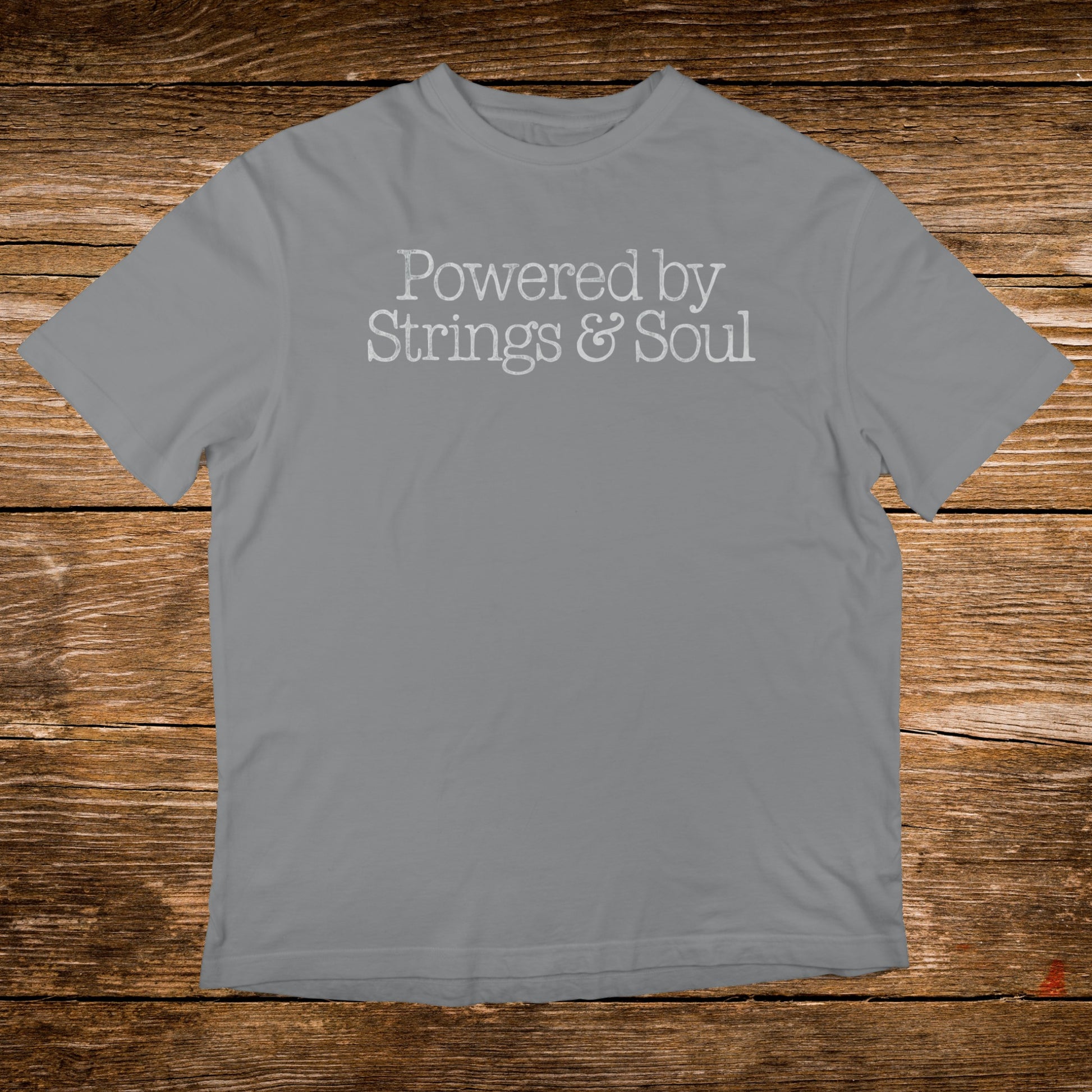 Grey t-shirt with "Powered by Strings & Soul" text on a wooden background.