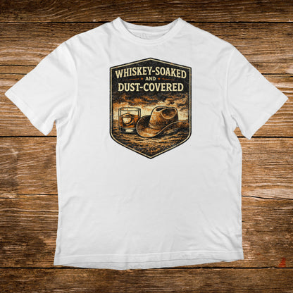 White t-shirt with "Whiskey-Soaked AND Dust-Covered" text and whiskey glass and hat illustration.