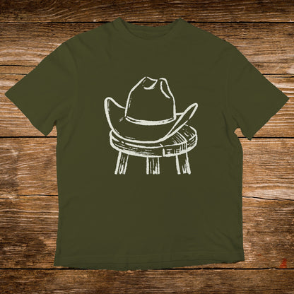 White cowboy hat rests on a wooden stool against olive green t-shirt.