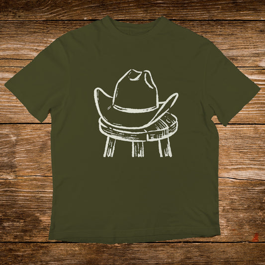 White cowboy hat rests on a wooden stool against olive green t-shirt.