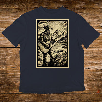 Man in cowboy hat plays guitar by river with mountains in background.